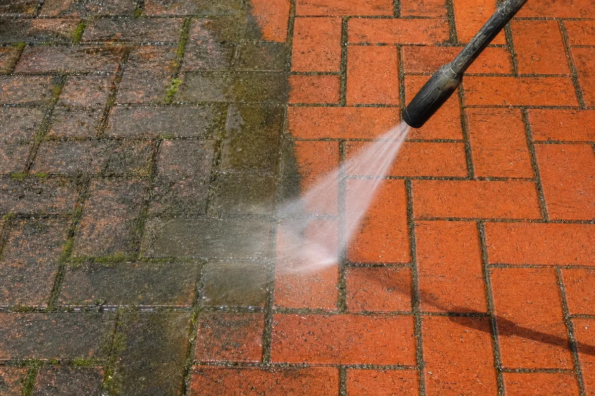 A red clay brick patio being pressure washed, with the cleaned half showing vibrant red brick next to the moss-covered dirty half